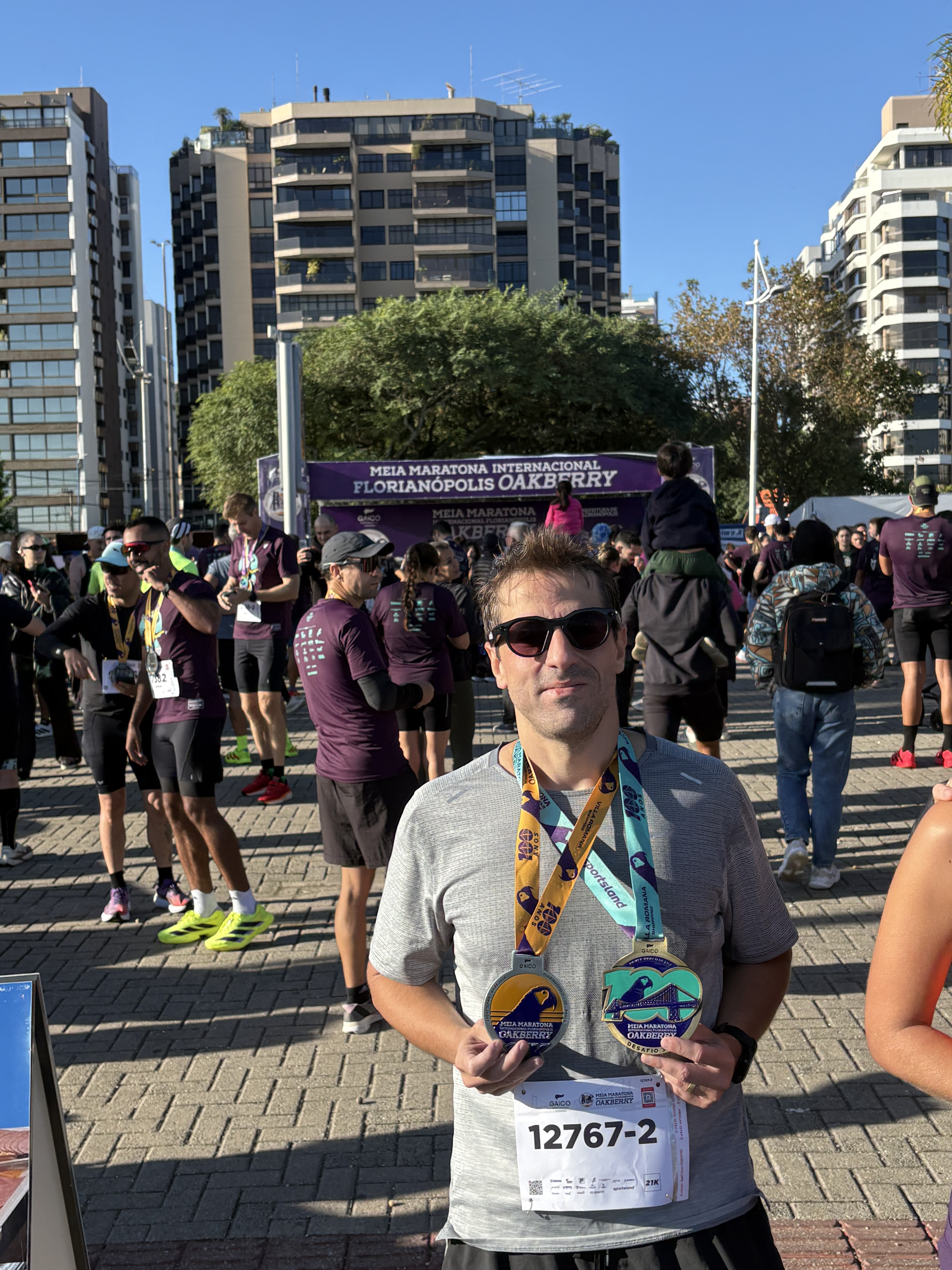 Atleta com duas medalhas da Meia Oakberry Floripa 2026 no peito, ambas com a Ponte Hercílio Luz estampada e o número 100 alusivo ao centenário, com a lona da 21ª edição da prova ao fundo