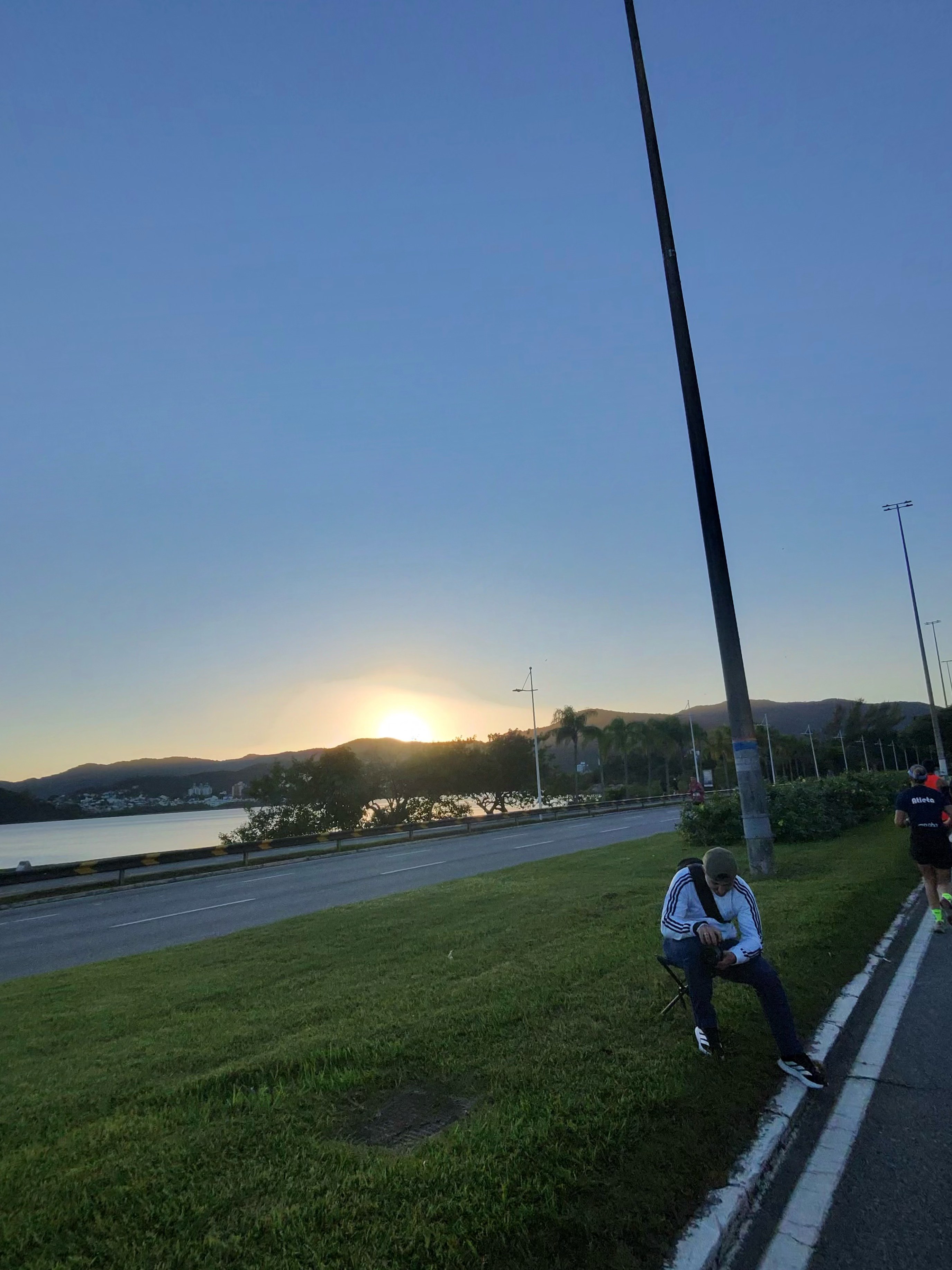 Sol nascendo atrás dos morros do continente visto da Avenida Beira-Mar Norte durante a Meia Oakberry, com baía calma refletindo a luz dourada e atleta passando à direita